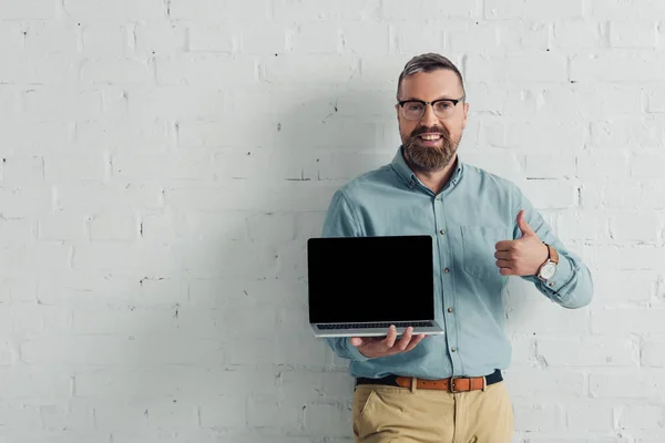 Guapo y sonriente hombre de negocios mostrando el pulgar hacia arriba y sosteniendo el ordenador portátil con espacio de copia - foto de stock