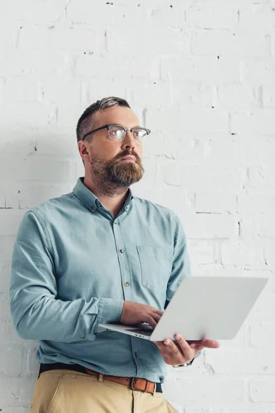 Homem de negócios bonito em camisa e óculos segurando laptop e olhando para longe — Fotografia de Stock
