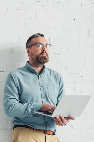 Bell'uomo d'affari in camicia e occhiali che tiene il computer portatile e distoglie lo sguardo — Foto stock