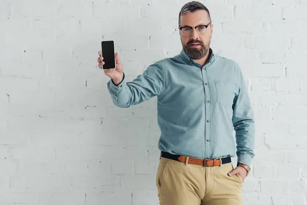 Hombre de negocios guapo con la mano en el bolsillo celebración de teléfono inteligente con espacio para copias - foto de stock