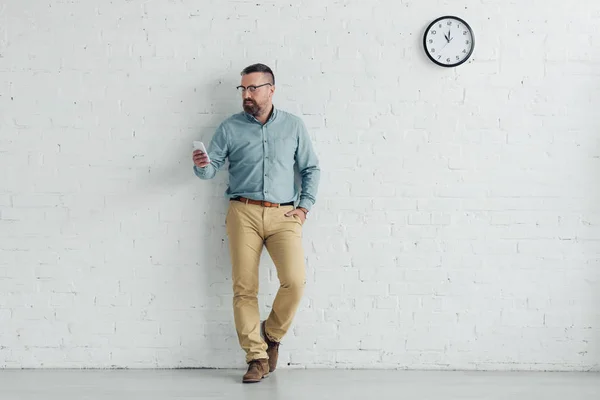 Handsome businessman with hand in pocket holding smartphone in office — Stock Photo