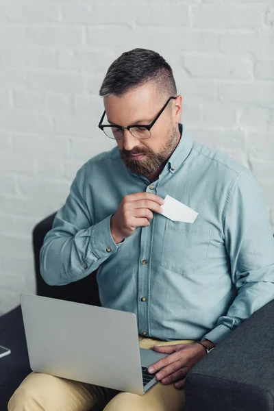 Bel homme d'affaires en chemise et lunettes tenant carte blanche et ordinateur portable — Photo de stock