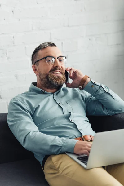 Handsome businessman in shirt holding laptop and talking on smartphone in office — Stock Photo
