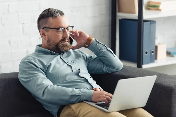 Bel homme d'affaires en chemise tenant ordinateur portable et parlant sur smartphone au bureau — Photo de stock