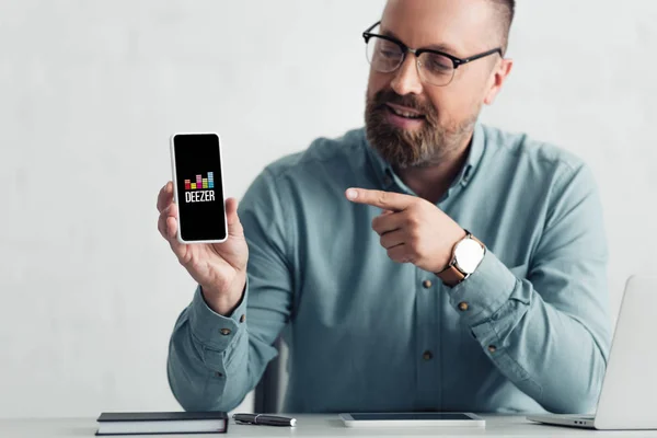 KYIV, UCRANIA - 27 de agosto de 2019: un hombre guapo con camisa que señala con el dedo al teléfono inteligente con el logotipo del deezer - foto de stock