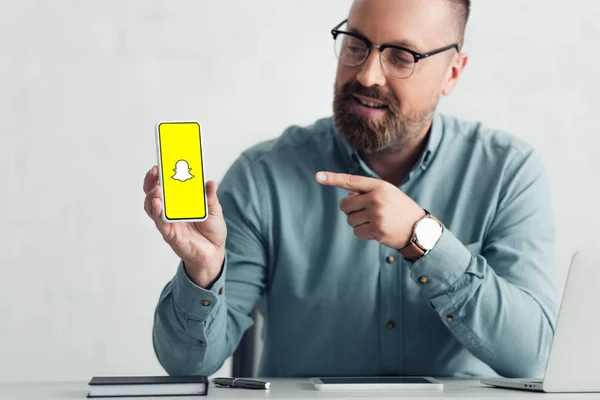 KYIV, UKRAINE - 27 de agosto de 2019: belo homem de negócios de camisa apontando com o dedo para o smartphone com logotipo Snapchat — Fotografia de Stock