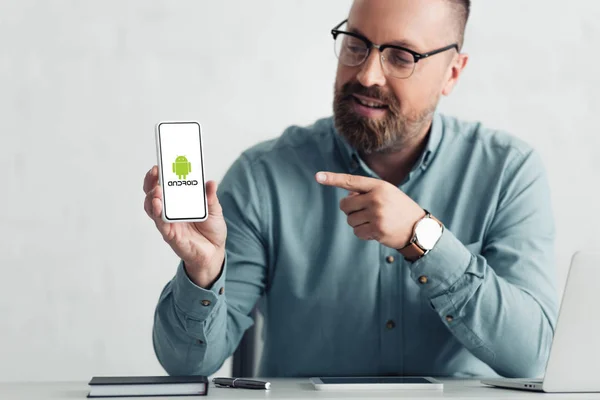 KYIV, UCRANIA - 27 de agosto de 2019: hombre de negocios guapo con camisa que señala con el dedo al teléfono inteligente con el logotipo de Android - foto de stock