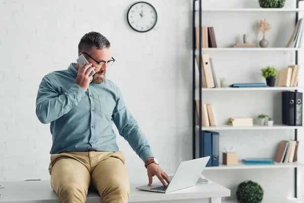 Homem de negócios bonito em camisa e óculos sentado na mesa e falando no smartphone — Fotografia de Stock