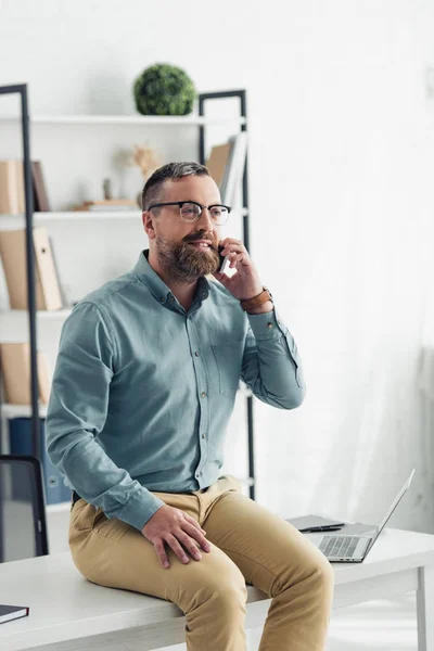 Красивый бизнесмен в рубашке и очках сидит на столе и разговаривает на смартфоне — стоковое фото