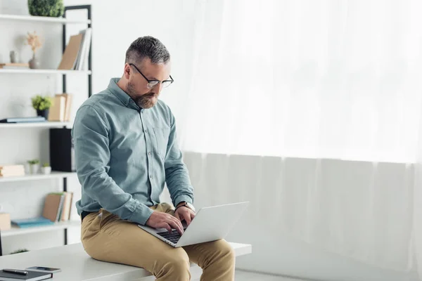 Homem de negócios bonito em camisa e óculos sentado na mesa e usando laptop — Fotografia de Stock