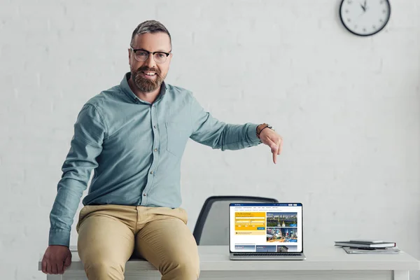 KYIV, UCRANIA - 27 de agosto de 2019: hombre de negocios guapo sentado en la mesa y apuntando con el dedo a la computadora portátil con el sitio web de reserva - foto de stock