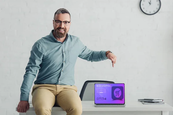 Schöner Geschäftsmann sitzt auf dem Tisch und zeigt mit dem Finger auf Laptop mit Web-Shopping-Website — Stockfoto