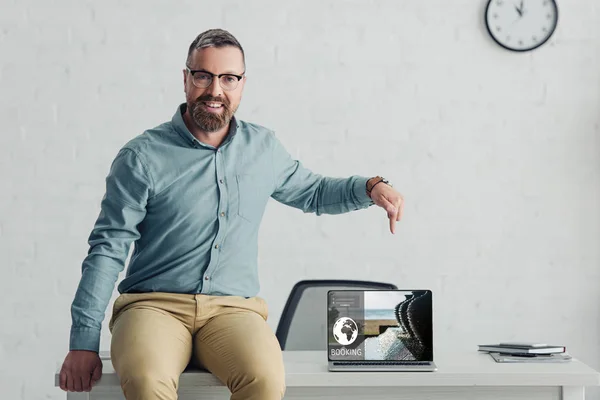 Homem de negócios bonito sentado na mesa e apontando com o dedo para laptop com site de reserva — Fotografia de Stock