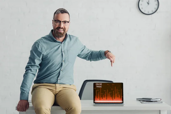 Guapo hombre de negocios sentado en la mesa y señalando con el dedo a la computadora portátil con el sitio web de comercio en línea - foto de stock