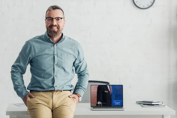 Schöner Geschäftsmann sitzt auf einem Tisch neben Laptop mit Buchungs-Website — Stockfoto