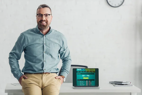 Bonito empresário sentado na mesa perto do laptop com site de apostas esportivas — Fotografia de Stock