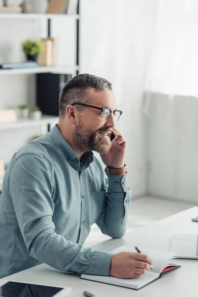 Bel homme d'affaires en chemise et lunettes parlant sur smartphone et tenant stylo — Photo de stock