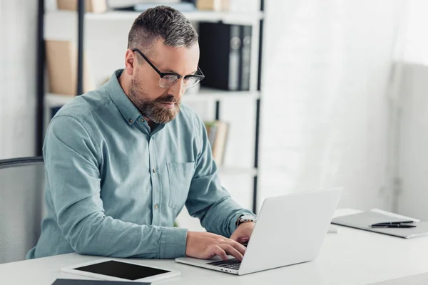 Homem de negócios bonito em camisa e óculos usando laptop no escritório — Fotografia de Stock