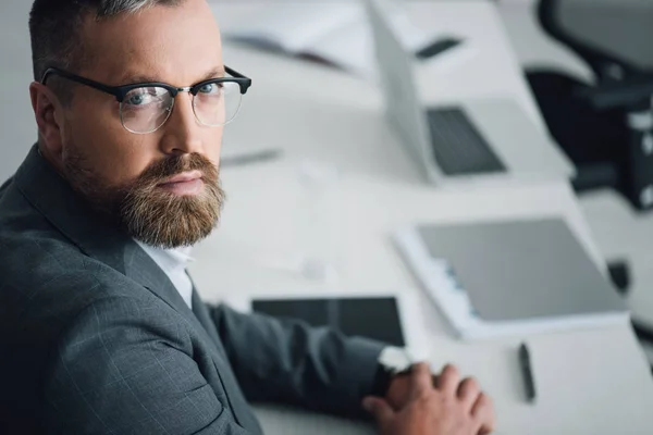 Guapo hombre de negocios en ropa formal y gafas mirando a la cámara en la oficina - foto de stock