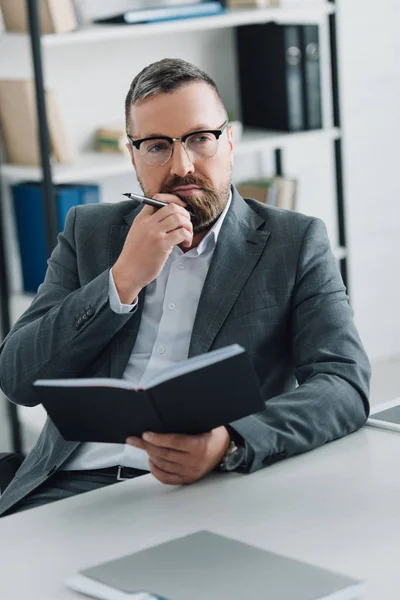 Красивый бизнесмен в формальной одежде и очках с ручкой и блокнотом в офисе — стоковое фото