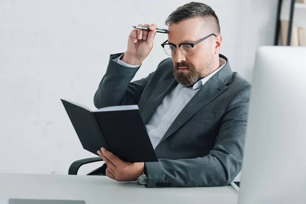 Bonito empresário no formal desgaste e óculos segurando caneta e notebook no escritório — Fotografia de Stock