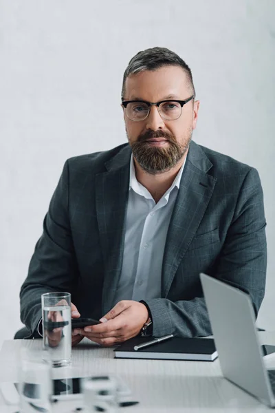 Homem de negócios bonito no desgaste formal e óculos segurando smartphone no escritório — Fotografia de Stock