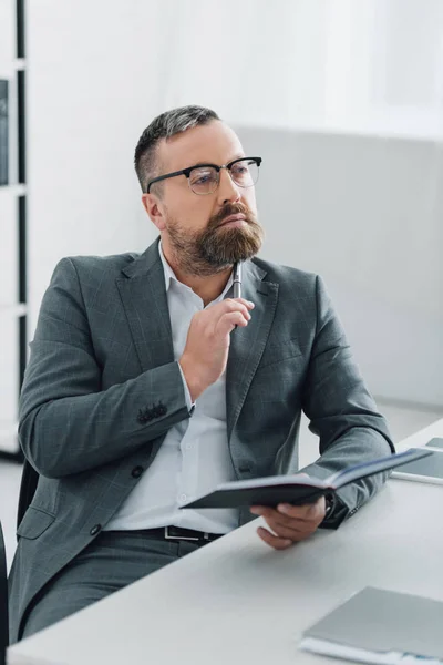 Schöner Geschäftsmann in formeller Kleidung und Brille mit Stift und Notizbuch im Büro — Stockfoto
