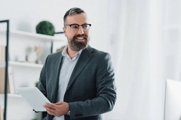 Guapo hombre de negocios en ropa formal y gafas usando tableta digital en la oficina - foto de stock