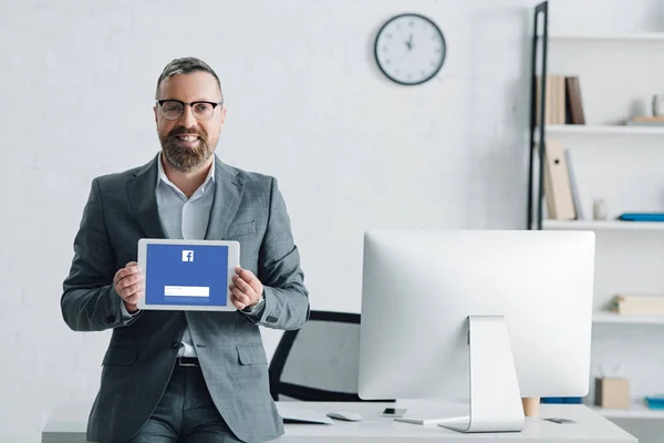 KYIV, UCRANIA - 27 de agosto de 2019: hombre de negocios guapo en ropa formal que sostiene la tableta digital con el sitio web de facebook - foto de stock