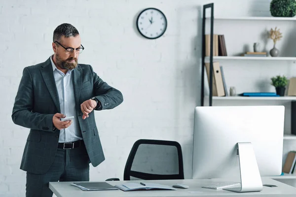 Handsome businessman in formal wear and glasses holding smartphone looking at watch — Stock Photo