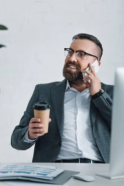 Guapo hombre de negocios en ropa formal y gafas hablando en el teléfono inteligente y sosteniendo la taza de papel - foto de stock