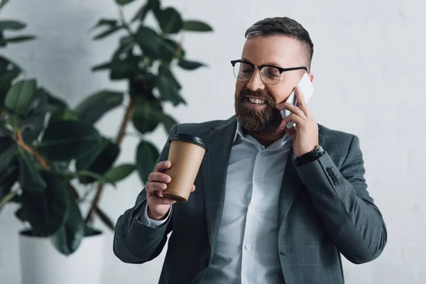 Handsome businessman in formal wear talking on smartphone and holding paper cup — Stock Photo