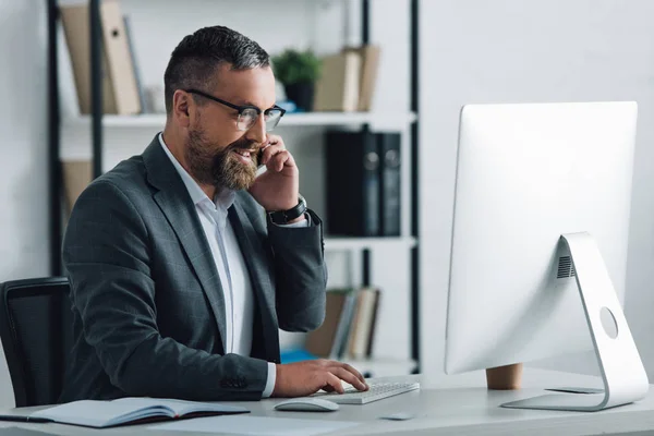 Hombre de negocios guapo en ropa formal hablando en el teléfono inteligente y mirando a la computadora — Stock Photo