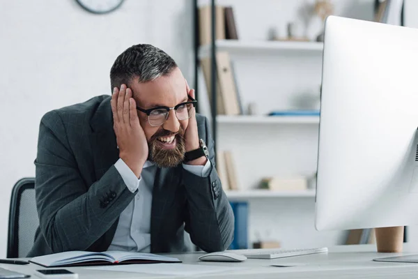 Hombre de negocios guapo en ropa formal hablando en el teléfono inteligente y mirando a la computadora - foto de stock