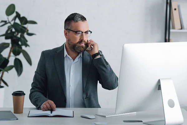 Schöner Geschäftsmann in formeller Kleidung, der auf dem Smartphone spricht und auf den Computer schaut — Stockfoto