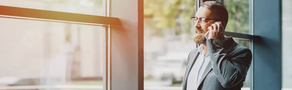 Panoramic shot of handsome businessman in formal wear and glasses talking on smartphone — Stock Photo