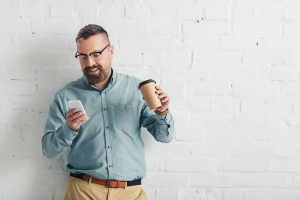 Guapo hombre de negocios en camisa y gafas sosteniendo teléfono inteligente y taza de papel - foto de stock