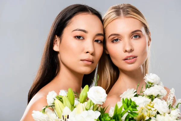 Beautiful blonde and asian brunette naked women with white flowers isolated on grey — Stock Photo
