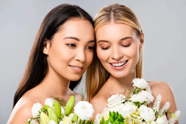 Smiling beautiful blonde and asian brunette naked women with flowers isolated on grey — Stock Photo