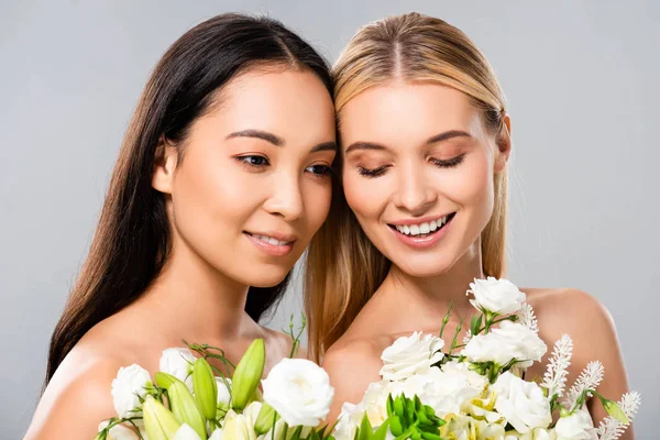 Sonriente hermosa rubia y asiática morena desnuda mujeres con flores aisladas en gris - foto de stock