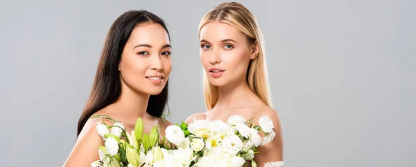 Smiling beautiful blonde and asian brunette naked women with flowers isolated on grey, panoramic shot — Stock Photo