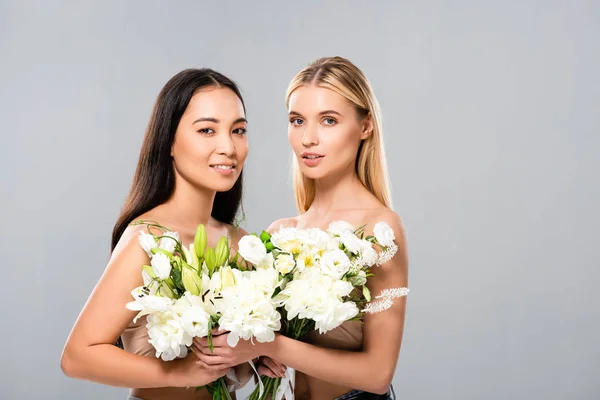 Smiling blonde and asian brunette naked women with flowers isolated on grey — Stock Photo