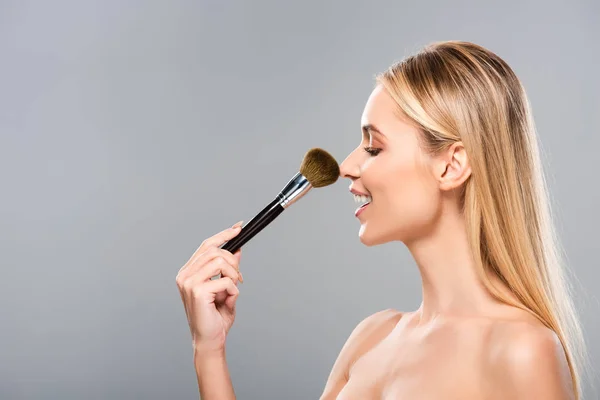 Seitenansicht der lächelnden jungen nackten Frau mit Kosmetikpinsel isoliert auf grau — Stockfoto