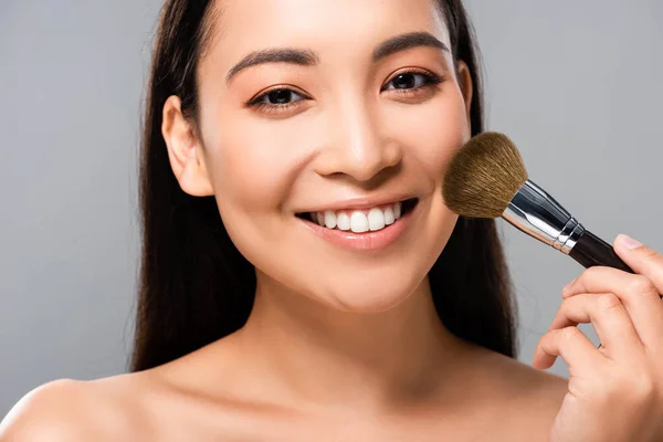 Sonriendo hermosa mujer asiática desnuda con cepillo cosmético aislado en gris - foto de stock