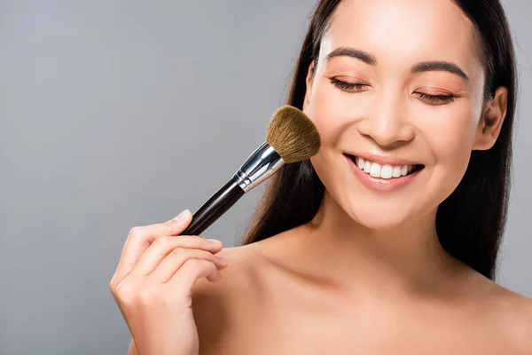 Happy naked asian woman with cosmetic brush isolated on grey — Stock Photo