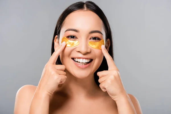 Sorrindo nu asiático mulher apontando com os dedos no olho remendos no rosto isolado no cinza — Fotografia de Stock