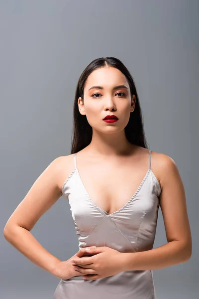 Привлекательная азиатка в атласном платье с красными губами, изолированными на сером — стоковое фото