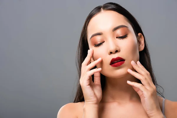 Hermosa mujer asiática con labios rojos tocando la cara aislado en gris - foto de stock