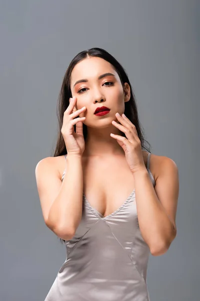Beautiful asian woman in satin dress with red lips touching face isolated on grey — Stock Photo