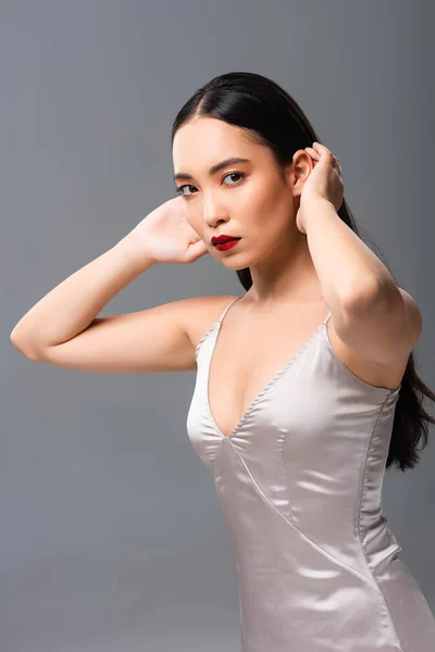 Beautiful asian woman in satin dress with red lips touching hair isolated on grey — Stock Photo